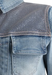 Jasnoniebieska kurtka dżinsowa z siateczkowym wzorem na ramionach, dwoma przednimi kieszeniami, kontrastującym przeszyciem i zaokrąglonym dołem.