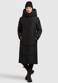 Abrigo acolchado largo negro con cuello alto y capucha, con secciones acolchadas, bolsillos laterales y una silueta recta. Combinado con botas negras.