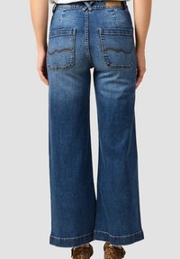 Szerokie niebieskie dżinsy z tylnymi kieszeniami, które mają falisty wzór przeszycia. Materiał ma gładką teksturę. Doły nogawek są surowe z widocznym przeszyciem.