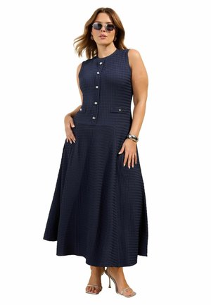Frau trägt ärmelloses, marineblaues, strukturiertes Midikleid mit silbernen Knöpfen, Sonnenbrille, Creolen, silbernem Armband und silbernen High-Heel-Sandalen.