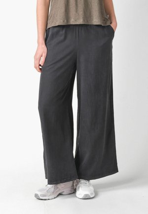 Személy laza fekete nadrágot és fehér tornacipőt visel, zsebre tett kézzel áll egy sima fehér háttér előtt.