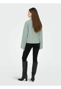Femme aux cheveux longs bruns portant une veste vert clair, un pantalon skinny noir et des bottes noires à talons montant jusqu'aux genoux, debout devant un fond uni.