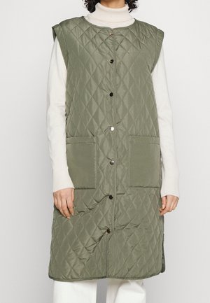 Bezrękawnik w kolorze oliwkowej zieleni, pikowany i długi, zapinany na zatrzaski z przodu, z dwoma dużymi kieszeniami, noszony na białym swetrze z golfem.