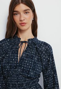 Kleid mit Marine-Muster, das über einen gerafften Ausschnitt, Bindeband und lange Ärmel verfügt. Der Stoff hat eine strukturierte Optik mit floralen Motiven.