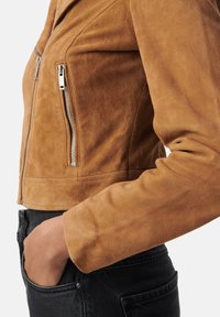 Vista lateral de una persona con una chaqueta de ante color beige con cremallera plateada y vaqueros negros, con la mano en el bolsillo.
