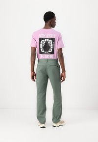 Rosa t-skjorte med svart grafisk design på ryggen, kombinert med grønne bukser og beige joggesko. Pent snitt og avslappet stil.