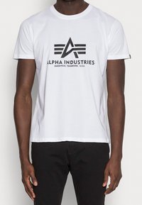 Bílý bavlněný tričko s kulatým výstřihem. Na přední straně se nachází černé grafické logo s nápisem "ALPHA INDUSTRIES" a horizontálními čarami.