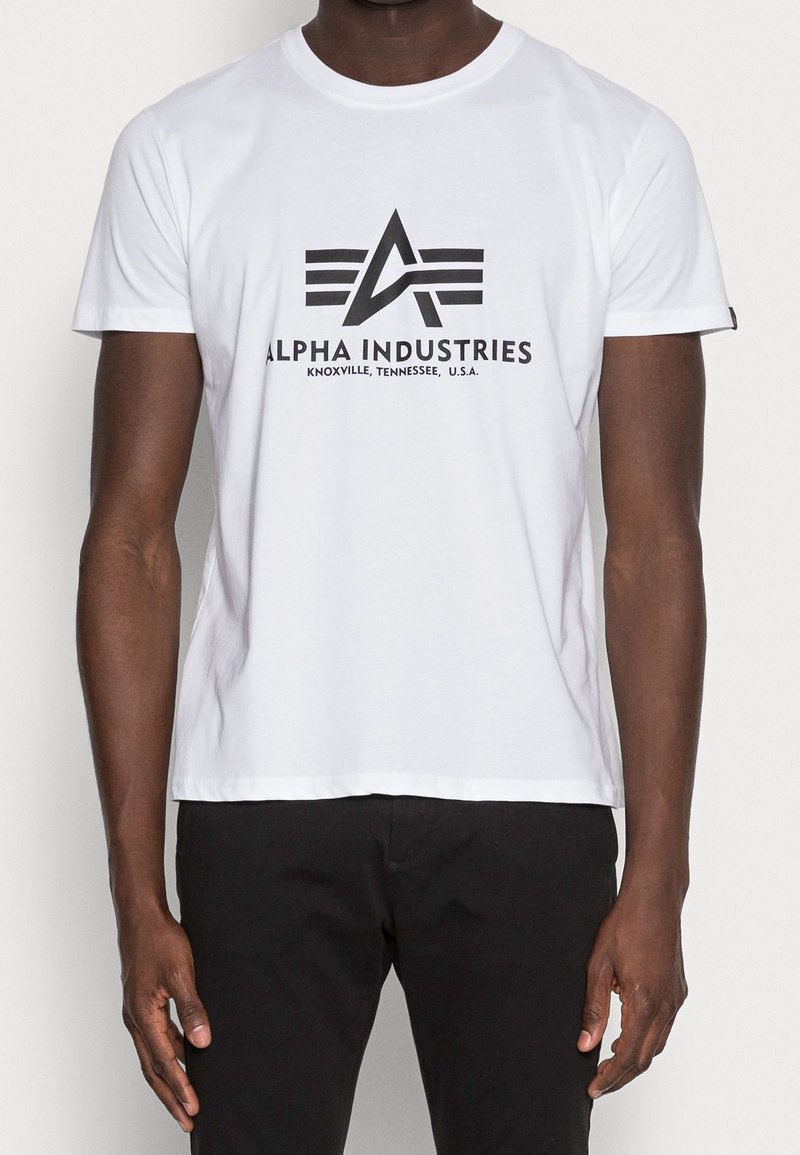 Bílý bavlněný tričko s kulatým výstřihem. Na přední straně se nachází černé grafické logo s nápisem "ALPHA INDUSTRIES" a horizontálními čarami.