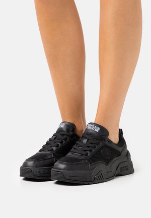 Czarne sneakersy wykonane z siateczkowych i skórzanych materiałów, z masywną podeszwą, sznurowanym zapięciem oraz logo na języku i boku. Widoczne szczegóły tekstury.