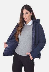 Giubbotto imbottito blu navy con zip frontale e cappuccio, caratterizzato da un interno trapuntato e due tasche laterali, indossato sopra un maglione grigio.