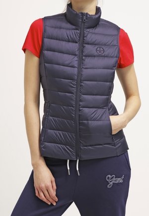 Femme portant un gilet matelassé bleu marine par-dessus un T-shirt rouge à manches courtes avec un pantalon de survêtement Gant bleu marine, la main dans la poche du gilet.