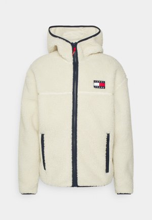 Kremowa fleecowa kurtka z kapturem, z suwakiem z przodu, bocznymi kieszeniami na suwak oraz naszywką z logo Tommy Jeans na piersi.
