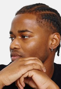 Portret van een persoon met gevlochten haar en een zwarte oorring, die hun kin op gekruiste vingers steunt, met een gladde huid en een ontspannen uitdrukking.