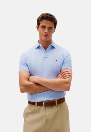 Hombre con cabello rizado de pie con los brazos cruzados, vistiendo una camisa polo azul claro y pantalones beige con un cinturón marrón.