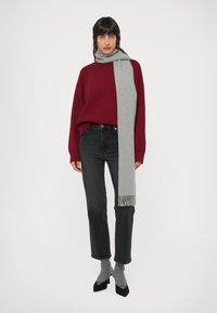 Μπορντό πουλόβερ, γκρι κασκόλ με φράγκα, μαύρα τζιν, γκρι κάλτσες και μαύρα τακούνια. Πρόσοψη μπροστά σε απλό λευκό φόντο.