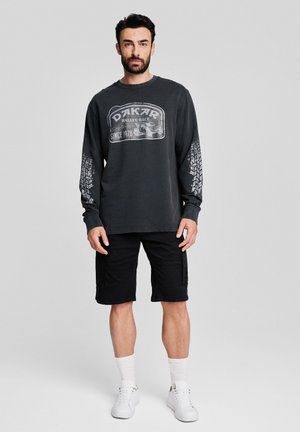 Czarna szara koszulka z długim rękawem z grafiką Dakar Rally, z białymi odciskami opon na rękawach, zestawiona z czarnymi szortami cargo i białymi trampkami.