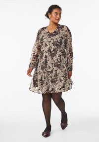 Abito corto a maniche lunghe con motivo floreale in tonalità beige, marrone e nero. Scollatura a V e gonna a strati. Abbinato a collant neri e scarpe borgogna.