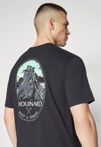 T-shirt en coton noir avec un motif d'une montagne et le texte "HOUINARD" et "CONÇU POUR DURER" au dos. Design à col rond.