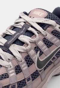 Sneaker Nike avec une tige en mesh bleu marine et rose pastel, des lacets blancs, un logo swoosh gris, et une étiquette "Huarache" sur la languette.