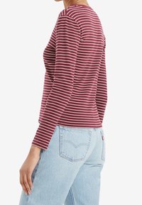 Maroon long-sleeve top with white horizontal stripes, featuring a round neckline and a fitted shape, paired with light blue denim jeans.