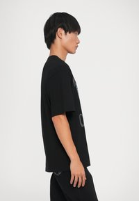 Μαύρο κοντομάνικο μπλουζάκι που προβάλλεται α�πό το πλάι, διαθέτοντας χαλαρή εφαρμογή και απλό σχέδιο. Λείο ύφασμα χωρίς ορατά μοτίβα.