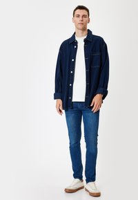 Camisa de mezclilla en azul oscuro, camiseta blanca debajo, jeans azules y zapatillas blancas con suela de goma marrón. Corte relajado y diseño informal.