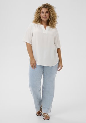 Vrouw met krullend haar die een witte losse top, lichtblauwe wijdbeen jeans en witte sandalen met bandjes draagt, staand voor een effen achtergrond.