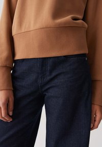 Persoon in een bruine trui met ribbels aan de zoom en manchetten, gecombineerd met donkerblauwe denim jeans, met ontspannen handen aan de zijkanten.