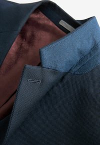 Marineblazer van duurzaam textiel. Blauw kraagmateriaal contrasteert met de binnenvoering in bordeauxrood met een geometrisch patroon. Netjes afgewerkte stiksels.