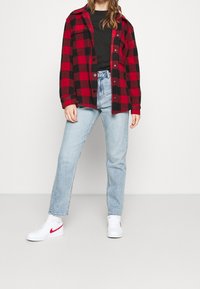 Chemise en flanelle à carreaux rouges et noirs, t-shirt noir en dessous, jean bleu clair à jambes droites, baskets blanches avec des accents rouges.