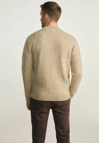 Beżowy sweter dzianinowy w łaty, okrągły dekolt oraz ściągacze na rękawach i dole. Nosi się go z ciemnobrązowymi spodniami, widok z tyłu.