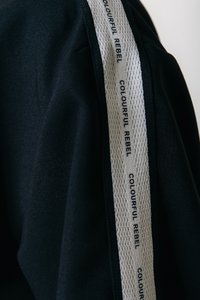 Sudadera con capucha negra de tela que presenta una franja lateral texturizada blanca con "COLOURFUL REBEL" impreso en negro. La vista de cerca resalta los detalles del material.