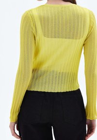 Kobieta ubrana w przezroczysty, żebrowany, żółty top z długim rękawem na czarną bieliznę i czarne spodnie, pokazana od tyłu na białym tle.