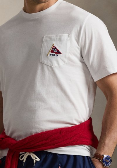 Άνδρας που φοράει λευκό μπλουζάκι με κόκκινο και μπλε λογότυπο Polo στην τσέπη, σκούρο μπλε σορτς με κορδόνι, κόκκινο πουλόβερ δεμένο γύρω από τη μέση και μπλε ρολόι στον καρπό.
