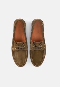 Ζευγάρι μοκασίνια από σουέντ σε ελιά πράσινο χρώμα με καφέ κορδόνια και λευκά ράμματα, πάνω σε λευκό φόντο, εμφανιζόμενα από πάνω.