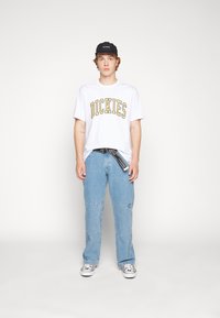 Hvitt T-skjorte med "DICKIES" i fet gul, kombinert med lyseblå cargo-bukser. Svart caps, svart belte og hvite sko med et mønster.