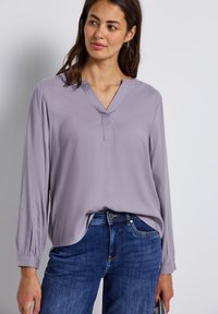 Blusa lunga a maniche con pizzo lavanda chiaro, scollatura a V, polsini plissettati e vestibilità ampia, abbinata a jeans in denim blu. Texture liscia.