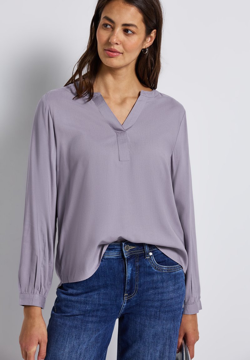 Blusa lunga a maniche con pizzo lavanda chiaro, scollatura a V, polsini plissettati e vestibilità ampia, abbinata a jeans in denim blu. Texture liscia.