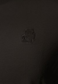 Petite silhouette noire brodée d'une personne avec une queue de cheval sur un fond de tissu foncé.