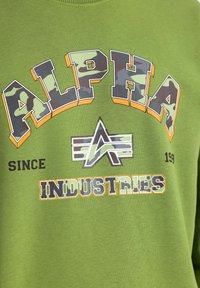 Felpa verde con la scritta "ALPHA INDUSTRIES" in caratteri in grassetto multicolori, con dettagli in camouflage e un emblema al centro.