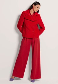 Roter doppelt knöpfiger Peacoat mit breiten Revers, kombiniert mit roten, weit geschnittenen Hosen. Das Model trägt lila Absätze und rote, runde Ohrringe.