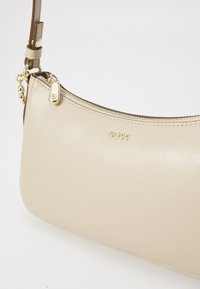 Sac à main en cuir beige avec une forme incurvée, doté d'une fermeture éclair et de quincaillerie dorées ; détail de logo embossé près de la fermeture éclair.