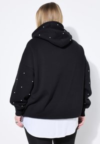 Sudadera negra con capucha y detalles de perlas en las mangas, corte relajado, y una camiseta blanca en capas visible en el dobladillo. Tejido suave y texturizado.