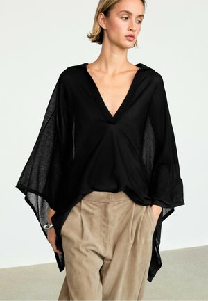Femme portant un haut noir fluide à col en V avec de larges manches et un pantalon beige taille haute, debout les mains dans les poches.