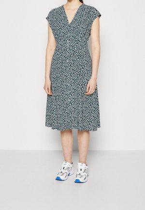 Femme portant une robe noire longueur genoux avec un imprimé floral blanc et des baskets blanches avec des touches bleues, debout devant un fond uni.