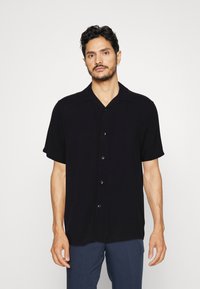 Chemise noire à manches courtes en tissu lisse, avec un devant boutonné, un col et une coupe décontractée, portée avec un pantalon bleu.