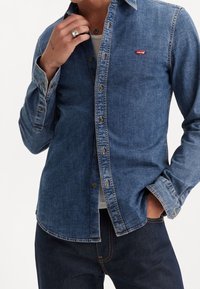 Camicia in denim di blu medio con colletto classico, chiusura frontale con bottoni e un piccolo logo rosso Levi's. Presenta maniche lunghe con bottoni ai polsini.