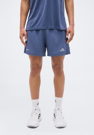 Άτομο που φορά μπλε αθλητικές σορτς Adidas, μπλε τοπ που ταιριάζει, λευκές κάλτσες Adidas και λευκά αθλητικά παπούτσια, στέκεται μπροστά σε απλό φόντο.