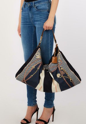 Mujer con vaqueros azules y tacones negros sostiene un gran bolso estampado con detalles de cadena y correa de cuero.