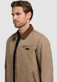 Homme aux cheveux bruns courts portant une veste beige avec un col marron foncé sur une chemise noire, tourné vers la gauche.